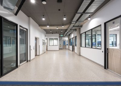 Bright modern office corridor with glass-walled rooms on both sides and a polished speckled floor, ceiling ducts and hanging lights visible in the aisle.