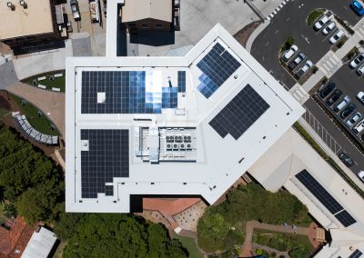 Aerial view of a large building complex with extensive solar panels on white roofs, adjacent parking lot, and surrounding greenery and roads.